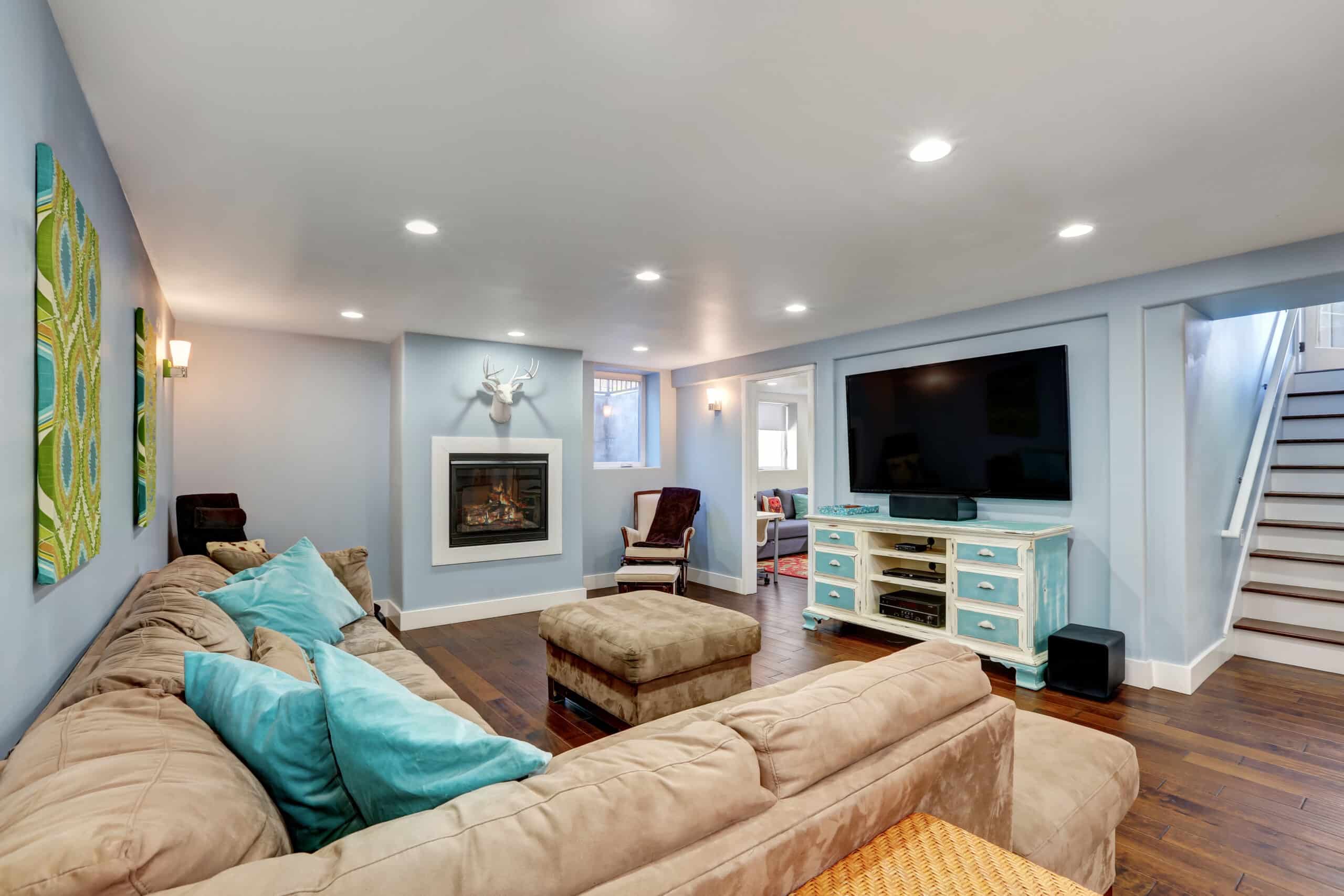 Pastel blue walls in basement living room interior. Large corner sofa with blue pillows and ottoman. Vintage white and blue TV cabinet.