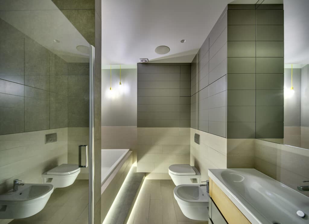 Bathroom in a modern style with tiles on the walls and floor. There is a sink with a faucet, white toilet and a bide, white bath, glass door, mirror, glowing lamps.