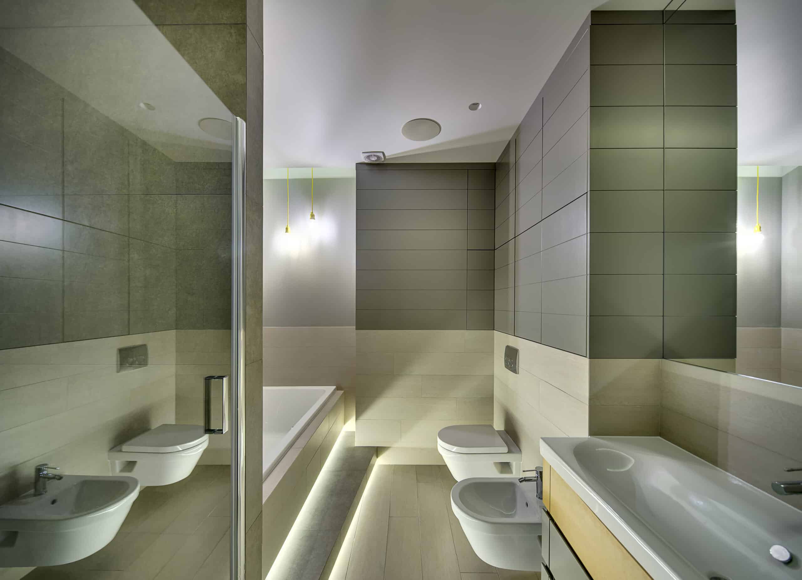 Bathroom in a modern style with tiles on the walls and floor. There is a sink with a faucet, white toilet and a bide, white bath, glass door, mirror, glowing lamps. Horizontal.