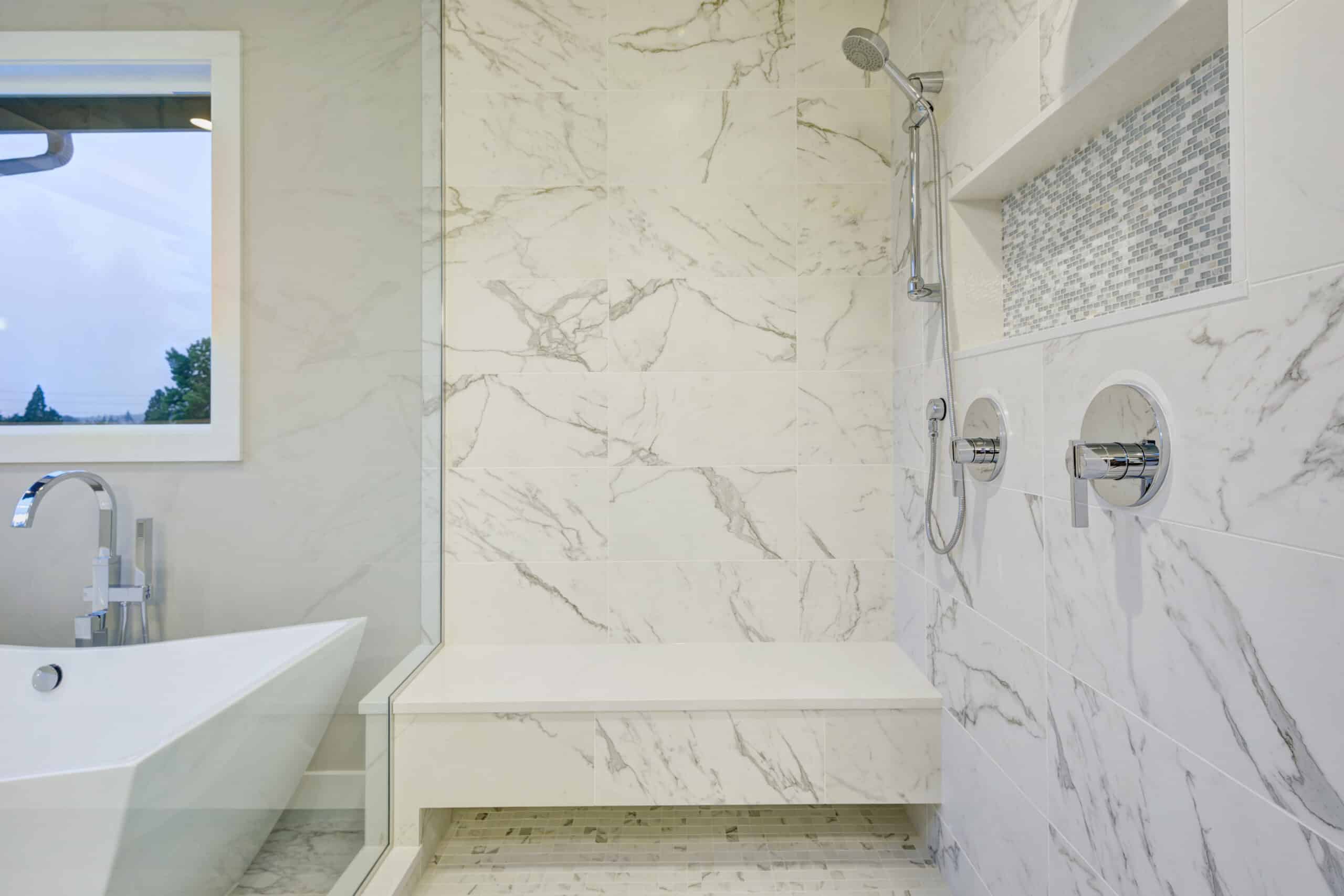 Sleek bathroom features freestanding bathtub and glass shower accented with rain shower head and gray and white marble surround.