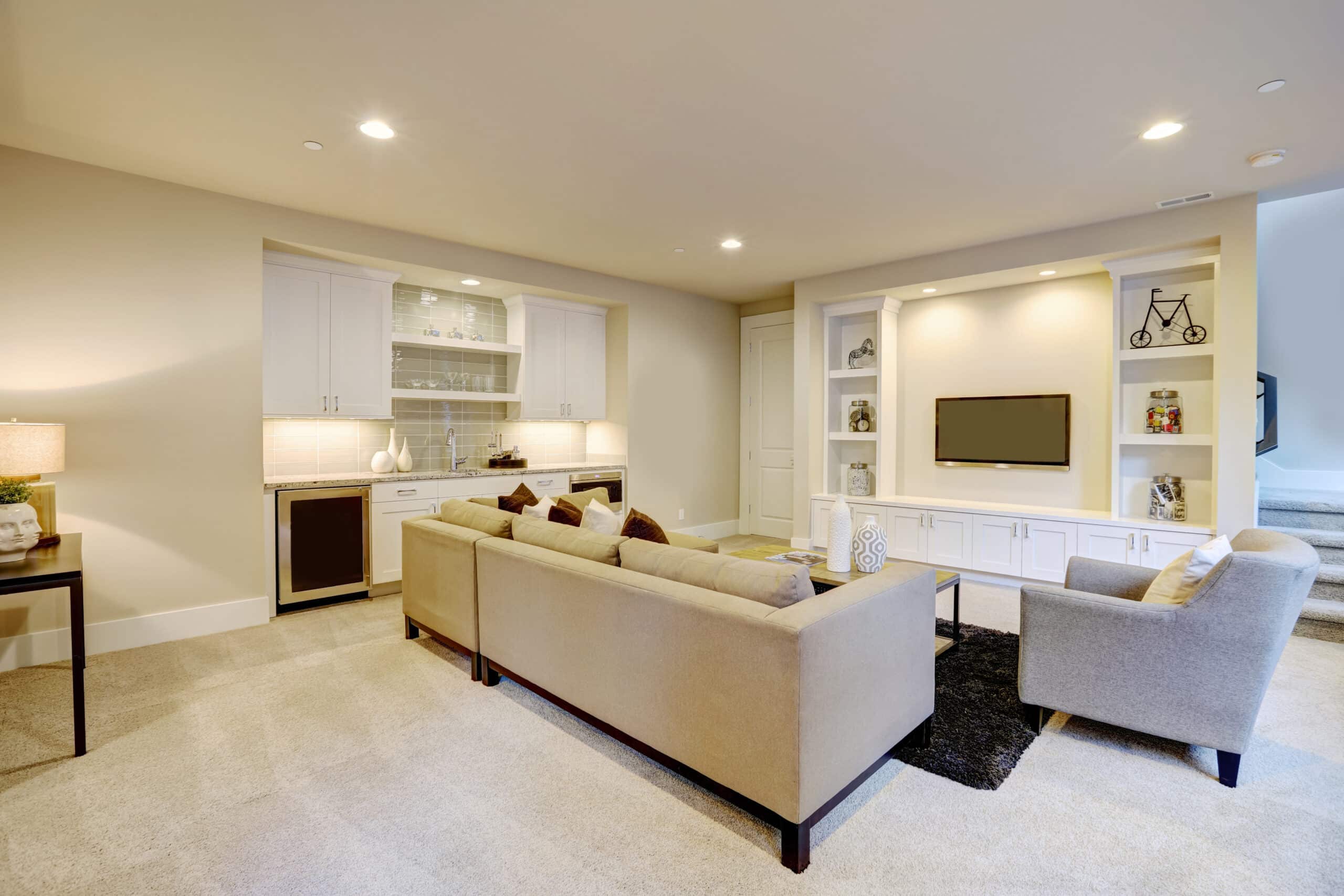 Chic basement features a gray sectional facing a white built-in tv cabinet and wet bar mounted to a wall.