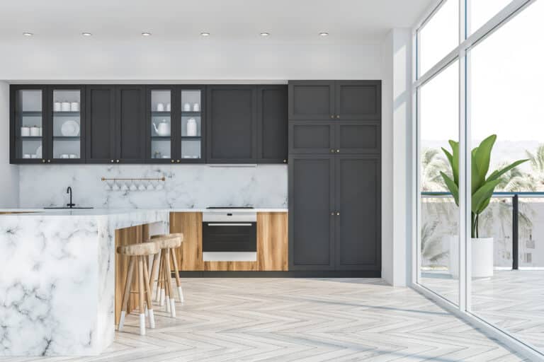 Interior of luxury kitchen with white and marble walls, wooden floor, large window with balcony, wooden countertops, gray cupboards and marble bar with stools.
